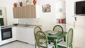 A compact kitchen with white cabinets, a built-in oven, and a dining area with green chairs and a table. - Residenza l' Ulivo (Parghelia)