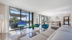 A modern living room with a large sofa, two armchairs, a coffee table, and a TV. There is a balcony with a view of the outdoors. - Luxurious Villa Amer with Private Pool in Bonaire Alcúdia (Alcúdia)