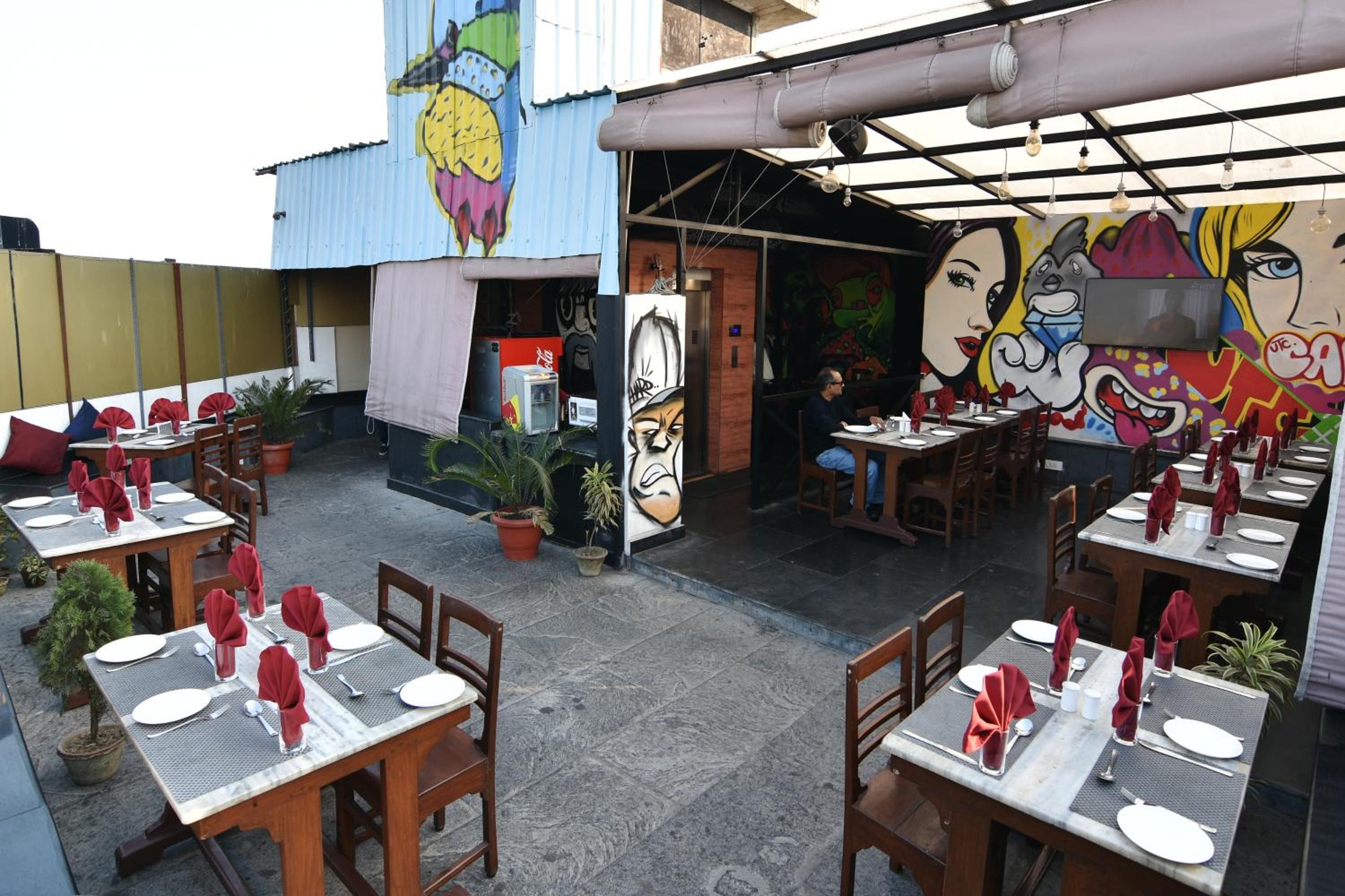 An outdoor dining area with tables set for two, potted plants, and a mural on the wall.