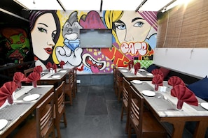 A restaurant interior with graffiti art on the walls, wooden tables set with red napkins, and a flat-screen TV mounted on the wall.