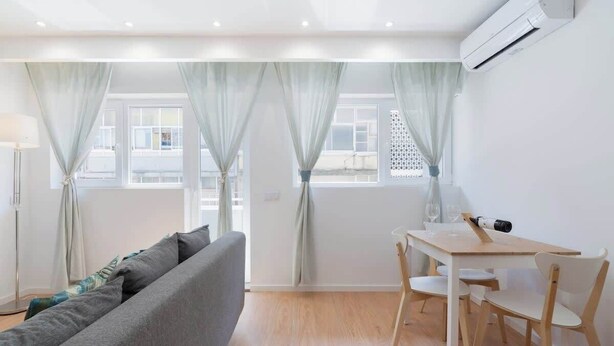 Una sala de estar moderna con un sofá gris, una mesa de comedor de madera con sillas y cortinas claras en las ventanas.