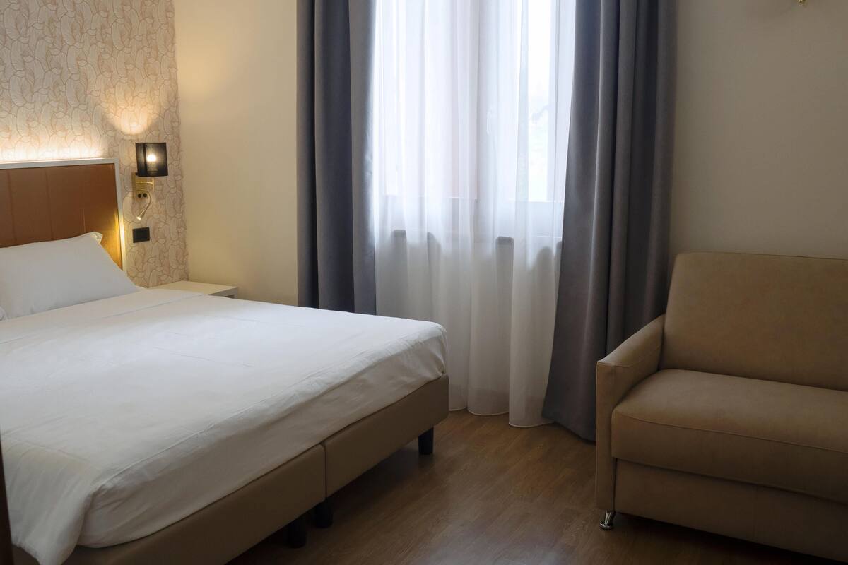 A hotel room with a bed, a sofa, a wall-mounted light, and a window with curtains.