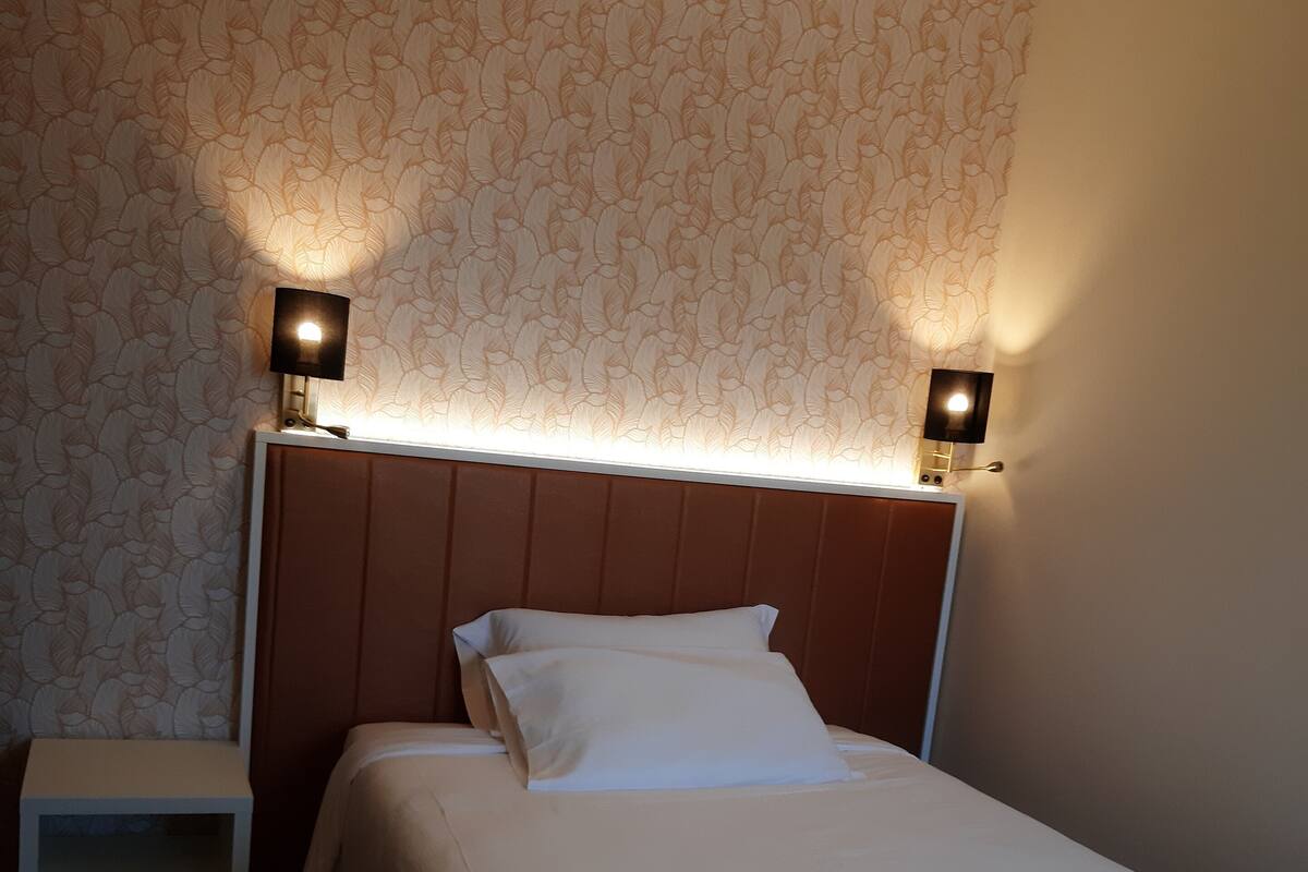 A neatly made bed with white linens in a room with patterned wallpaper and two wall-mounted lamps.