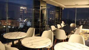 A high-rise hotel room with a city view, featuring a dining area with round tables and chairs, and a large window offering panoramic views. - Bigland Bogor Hotel Powered by Archipelago (Bogor)