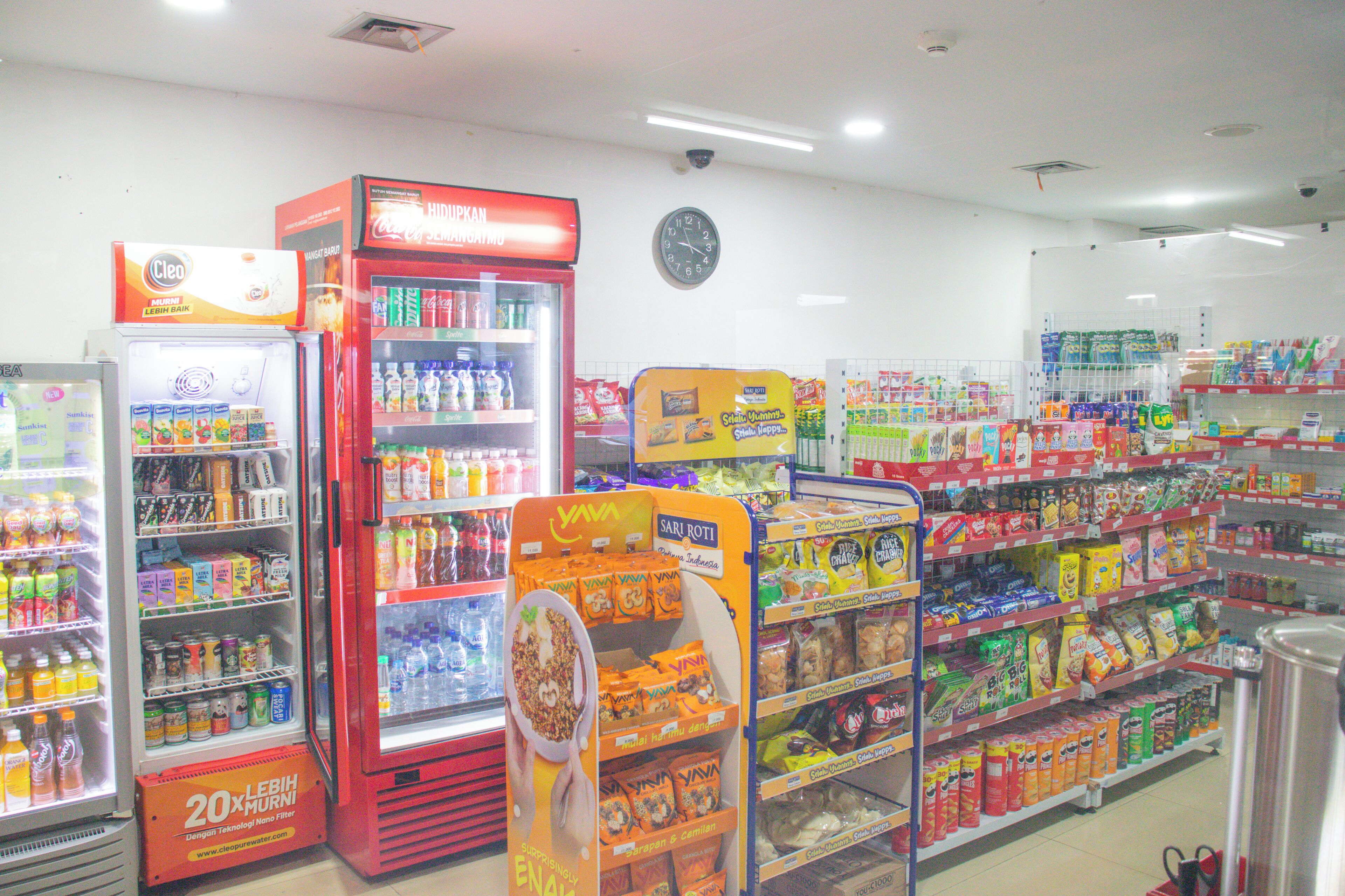 A grocery store with refrigerated sections, shelves stocked with snacks, and a visible clock on the wall.