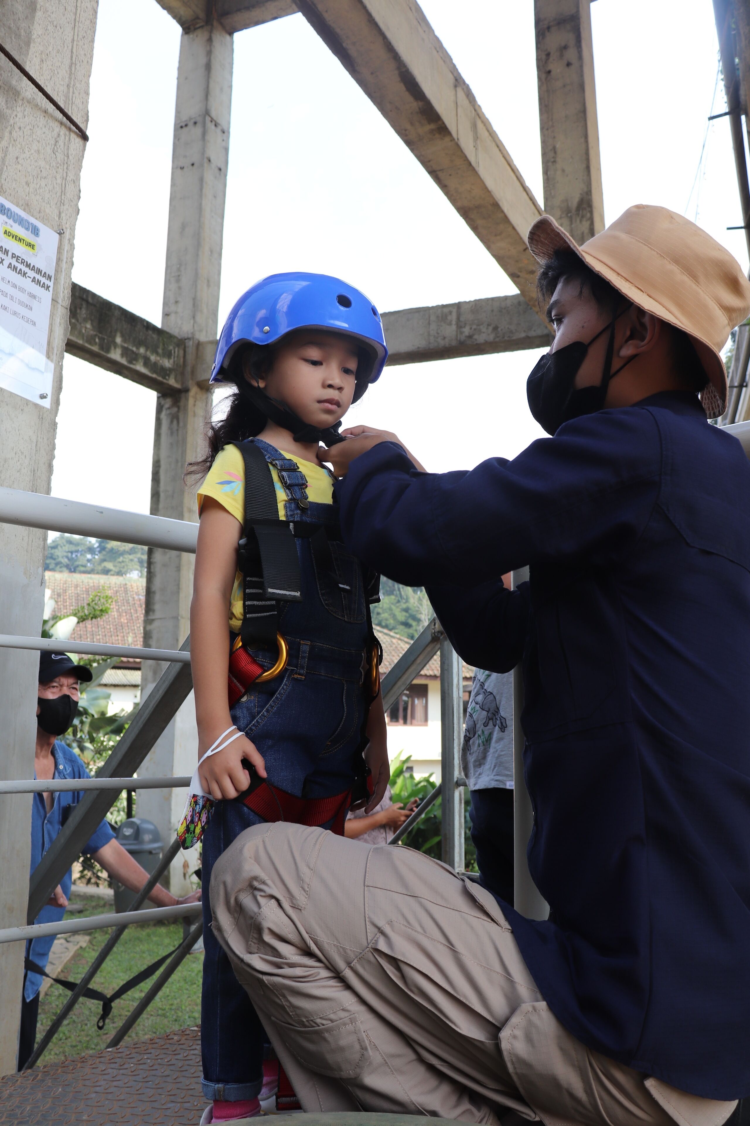Una persona con camisa azul y pantalones beige ayudando a un niño con casco azul y arnés de seguridad.