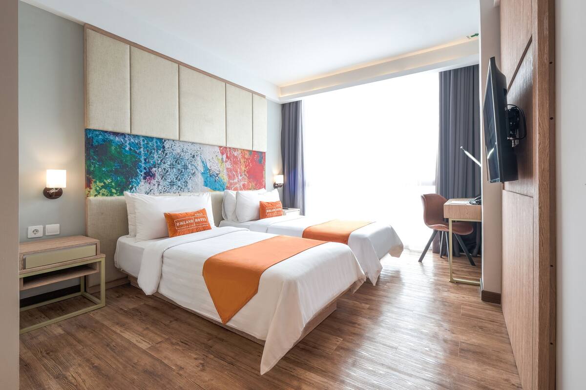 A hotel room with two beds, a desk, a TV, and a colorful abstract painting on the wall.