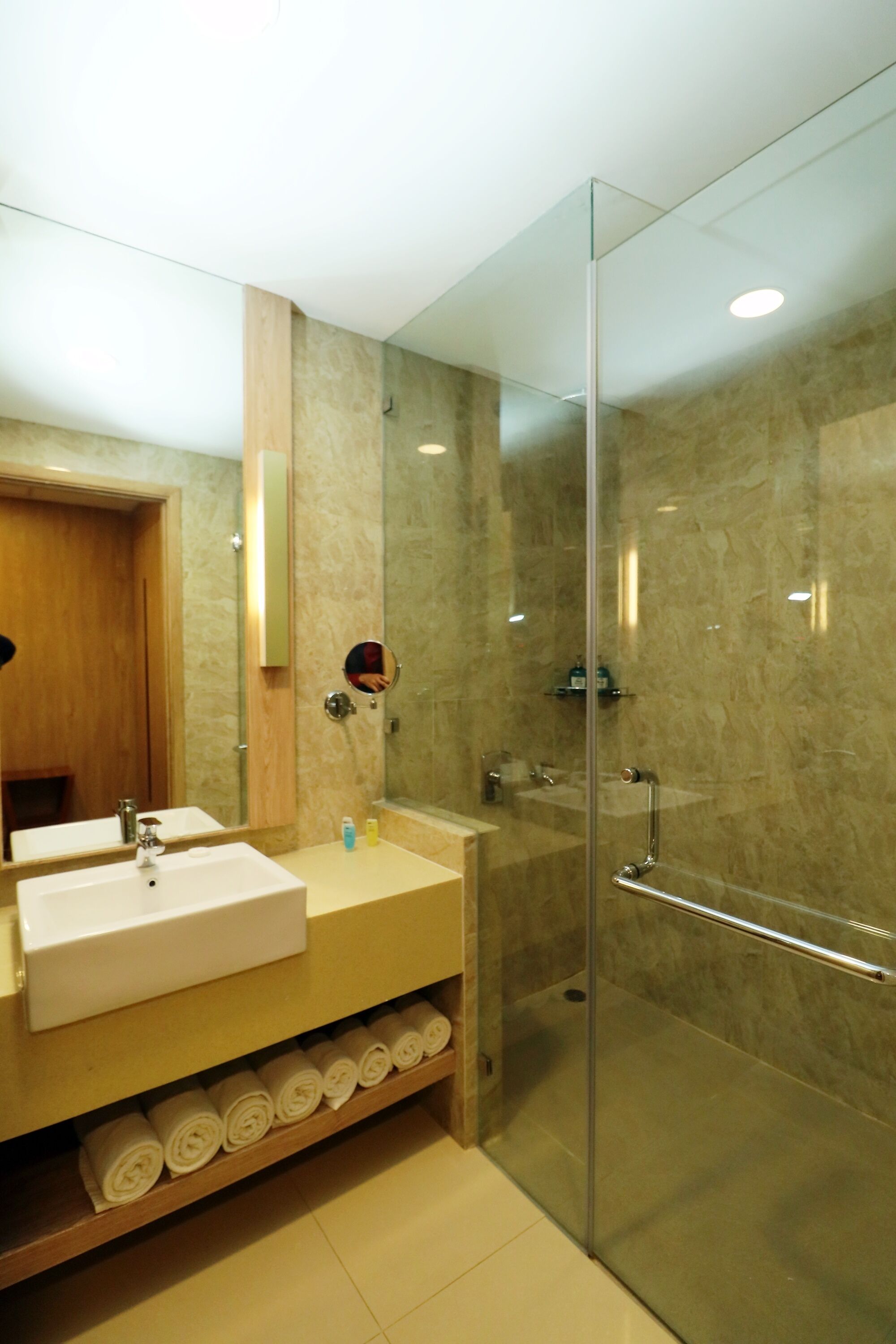 A modern bathroom with a glass-enclosed shower, a wooden vanity with a mirror, and a shelf with rolled towels.