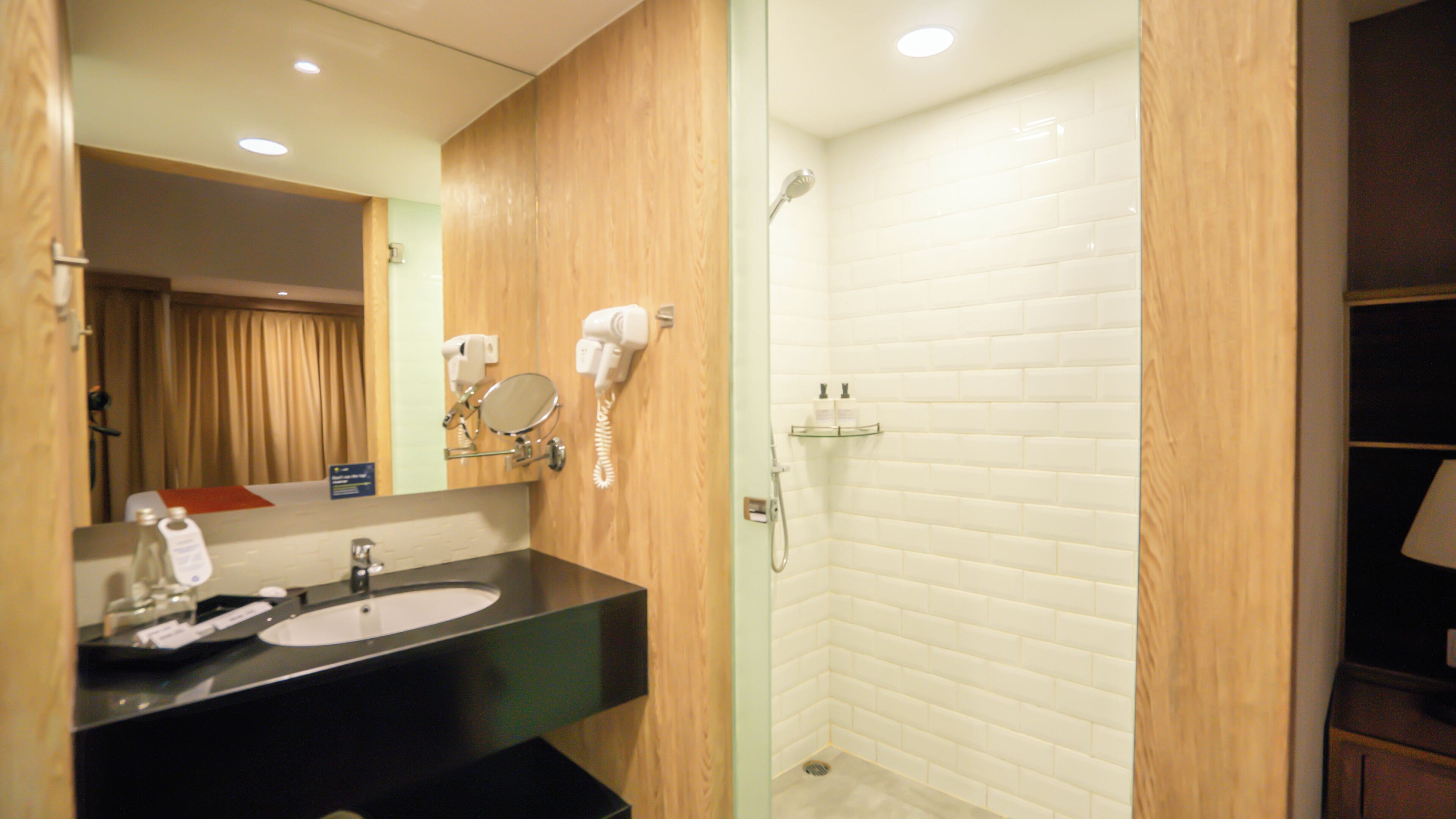 A hotel bathroom with a wooden vanity, a sink, a mirror, a hairdryer, and a shower with white tiles.