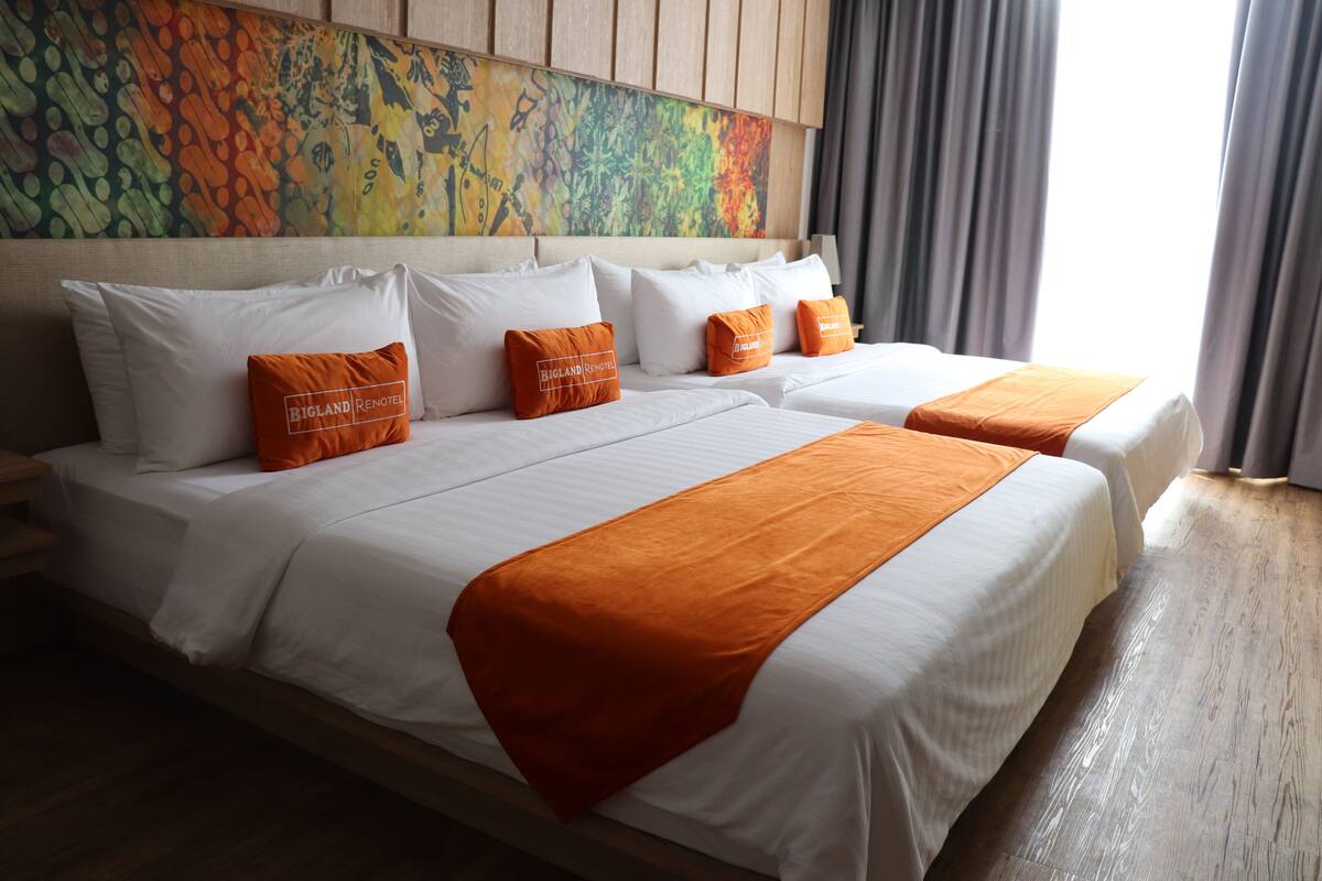 A hotel room with two beds, white bedding, orange throw pillows, and a colorful abstract painting on the headboard.