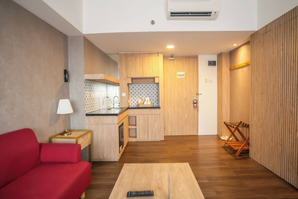 A compact living space with a red sofa, a small table, a kitchen area with wooden cabinets, and a wooden floor.