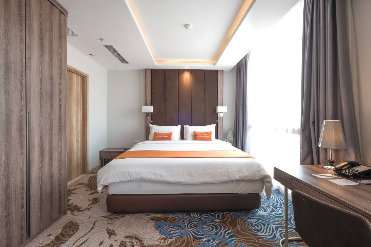 A hotel room with a large bed, wooden wardrobe, desk with a lamp, and a view of the outside.