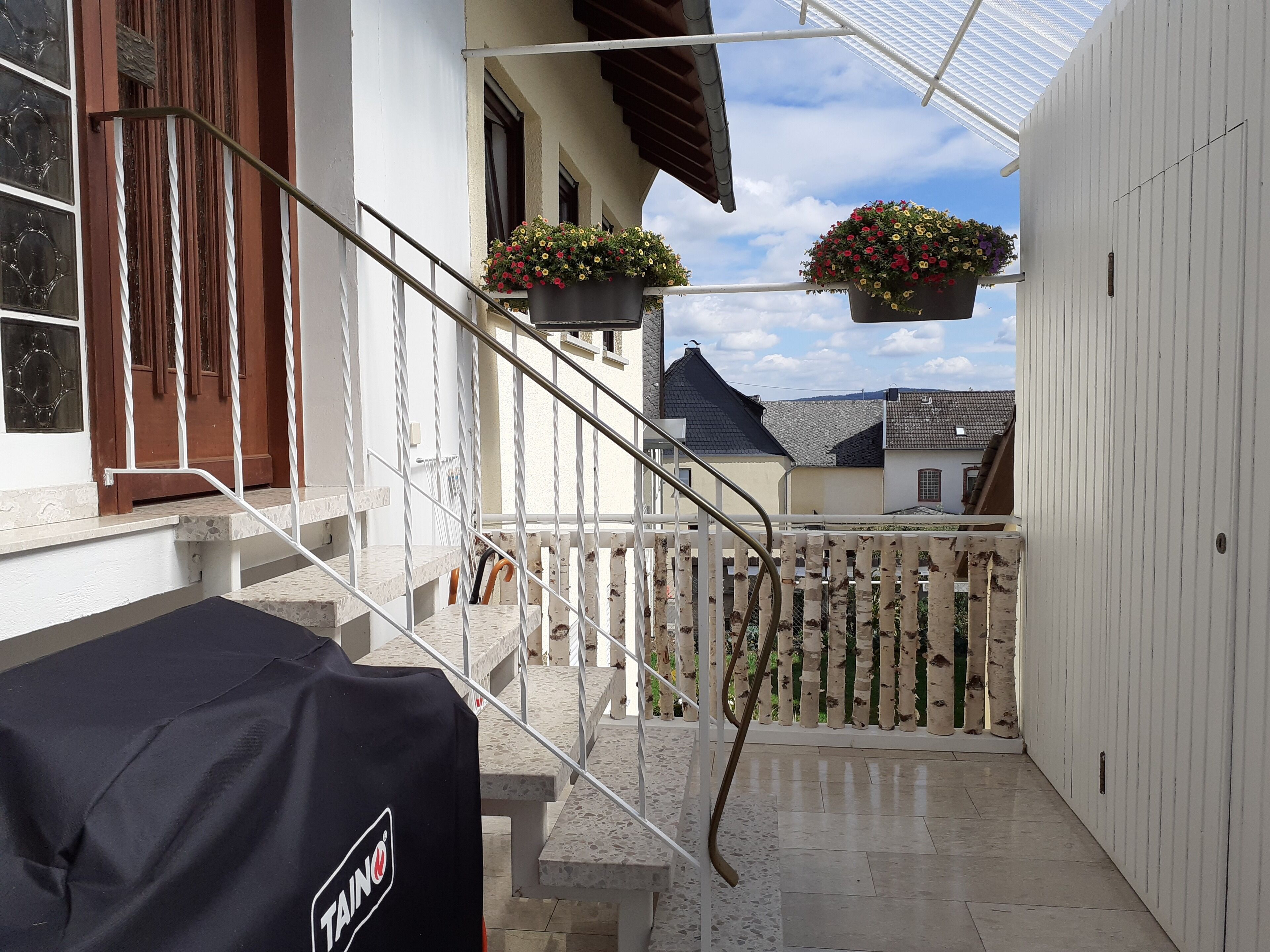 A covered outdoor area with a staircase leading to a balcony, a covered grill, and hanging flower baskets.