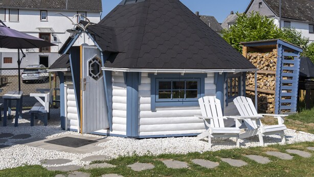 En timmerstuga med ett svart tak, vita väggar och en liten veranda med två vita Adirondack-stolar.