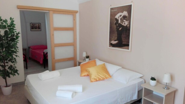 Ein Schlafzimmer mit Bett, Kissen, Nachttisch, Lampe, einem Blumentopf, einer Schiebetür und einem gerahmten Bild an der Wand.