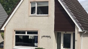 Une maison à deux étages, avec une façade beige, un toit marron et une porte blanche. Une fenêtre donne sur la rue et il y a un petit jardin avec des plantes.