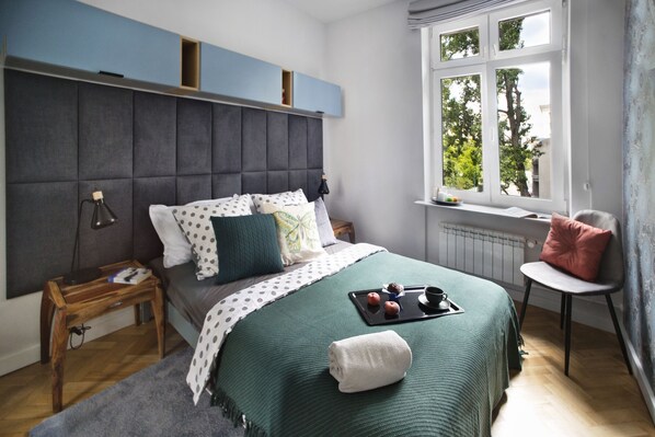 A bedroom with a bed, a nightstand, a chair, a window with a view of trees, and a radiator. - P&O Apartments Siewierska (Warsaw)