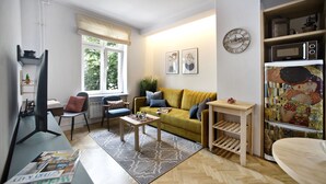 A modern living room with a sofa, coffee table, and a TV. There is a window with a view of trees, a clock on the wall, and a microwave on a shelf. - P&O Apartments Siewierska (Warsaw)