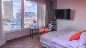 Habitación de hotel con un ventanal grande, una cama, una silla, una mesita y un televisor.