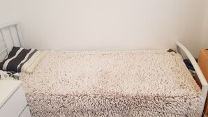 A small, neatly made bed with a fluffy white cover, a white bedside table, and a small wooden nightstand with a lamp.