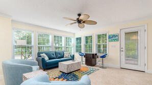 A spacious living room with a blue sofa, two armchairs, a coffee table, and a ceiling fan. Large windows allow natural light to fill the room. - FANTASY ISLAND LIFE (Jensen Beach)