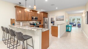 A modern kitchen with a central island, bar stools, wooden cabinets, stainless steel appliances, pendant lighting, and a view into the living area. - FANTASY ISLAND LIFE (Jensen Beach)