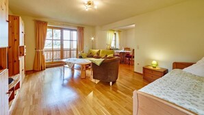 A spacious room with a wooden floor, a bed, a desk, and a chair. There is a window with curtains and a door on the left side.