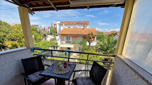 Uma varanda com mesa e duas cadeiras, com vista para uma área residencial com casas e vegetação.