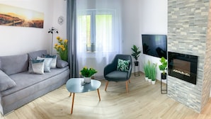 Una sala moderna con un sofá gris, un sillón negro, una pequeña mesa de centro, un televisor de pantalla plana, una chimenea y un amplio ventanal con cortinas.