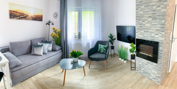 A modern living room with a grey sofa, a black armchair, a small coffee table, a flat-screen TV, a fireplace, and a large window with curtains.