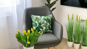 Un salón moderno con un sillón gris, una mesa de centro redonda de vidrio y una planta en maceta con flores amarillas.