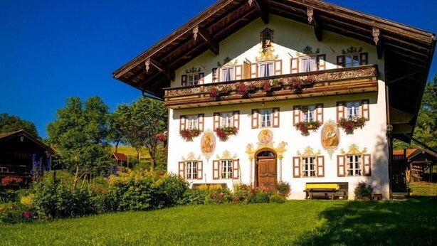 A traditional Alpine-style building with a sloped roof, wooden balconies with flower boxes, and a central entrance with a door. - Ferienwohnung Heuberg mit 1 Separaten Schlafzimmer (Samerberg)