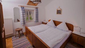 A bedroom with a wooden bed, a nightstand, a chair, a window with curtains, a radiator, and a ceiling light fixture. - Ferienwohnung Heuberg mit 1 Separaten Schlafzimmer (Samerberg)