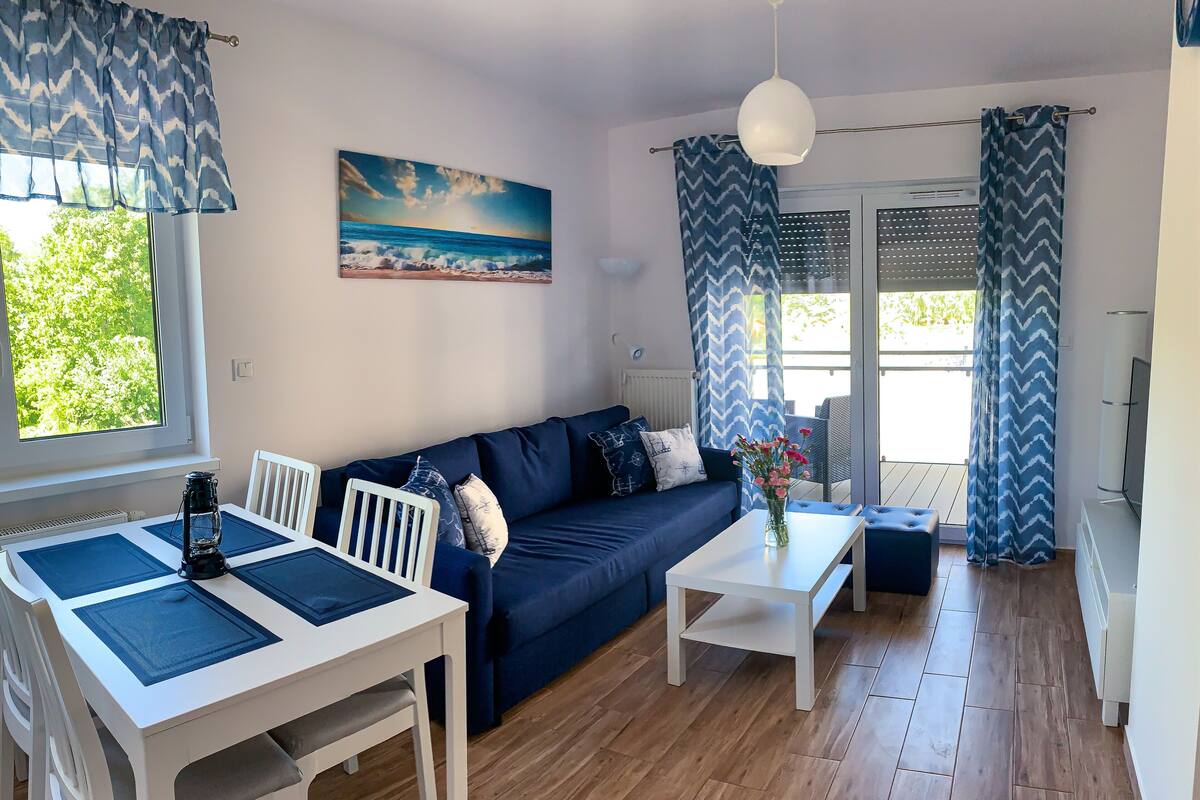 Un salon moderne comprenant un canapé, une table à manger et des chaises, un tableau représentant une scène de plage et de grandes fenêtres avec des rideaux.