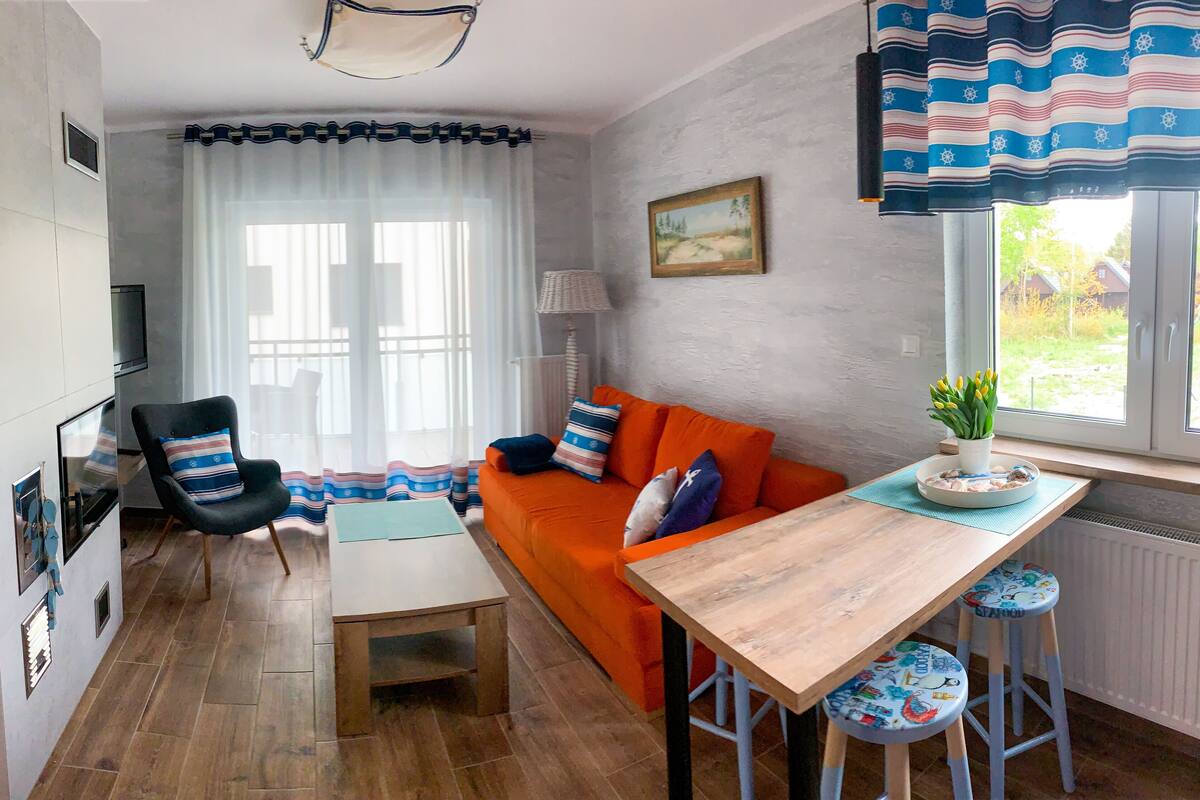 Un salon chaleureux avec une table en bois, un canapé orange et un fauteuil. On y trouve une fenêtre avec des rideaux, une lampe et une petite plante sur le rebord de la fenêtre.