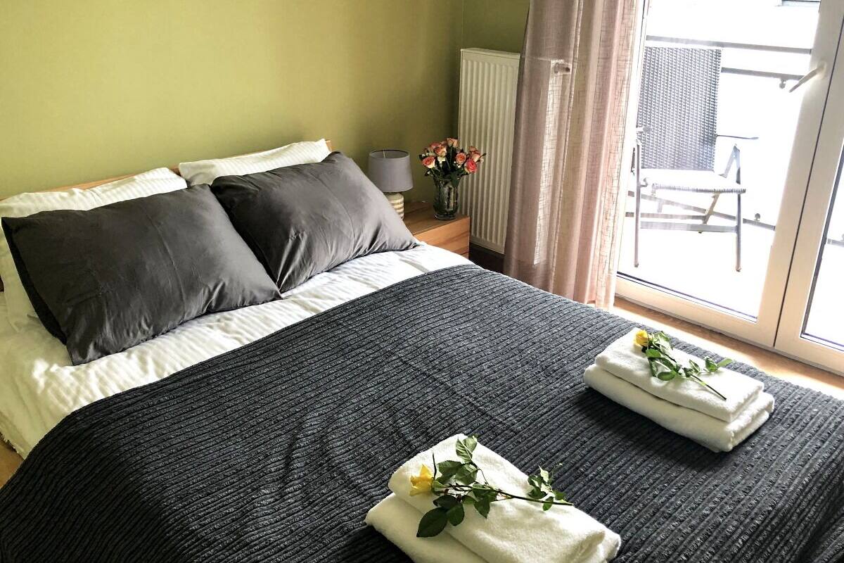 Un lit bien fait, recouvert d’une couverture et de coussins gris foncé, un vase rempli de fleurs posé sur une table de chevet, et une vue sur l’extérieur par une fenêtre.