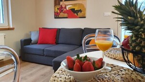 A living room with a sofa, a bowl of strawberries, a glass of orange juice, and a pineapple on a woven mat. - Apartamenty Szafirowa Mierzeja - 365PAM (Mielno)