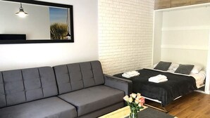 Moderni huone, jossa on harmaa sohva, puinen sohvapöytä, jossa on maljakko täynnä kukkia, mustalla peitteellä varustettu sänky ja valkoinen tiiliseinä.