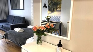 Hotellihuone, jossa on sänky, yöpöytä maljakko täynnä kukkia, pullo, peili ja ikkunasta avautuva näkymä rannalle.