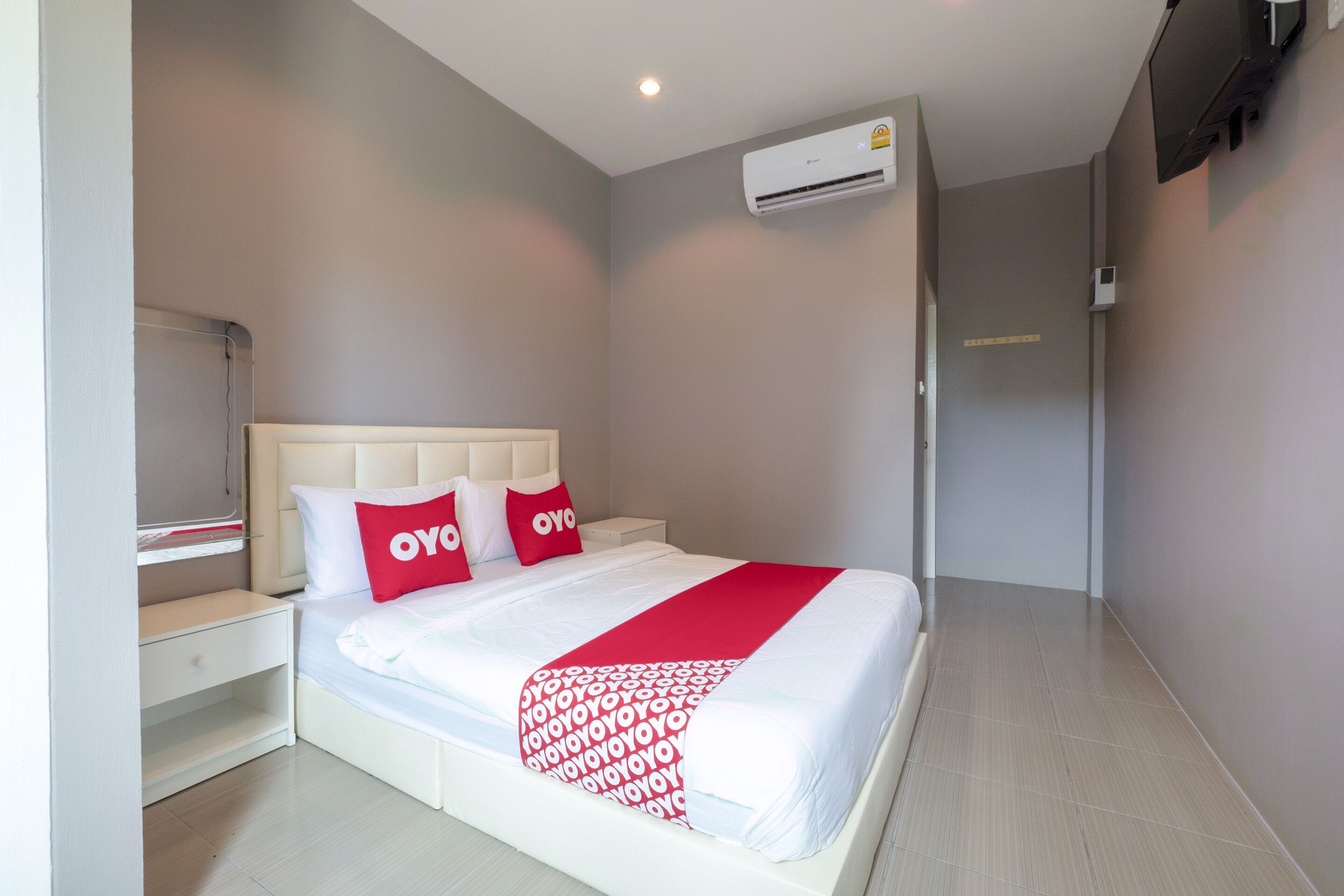 Un lit bien fait, avec une tête de lit blanche, deux oreillers ornés du logo « OYO » et une couverture rouge. On aperçoit une table de chevet blanche à côté du lit, un climatiseur fixé au mur et un téléviseur monté sur le mur.