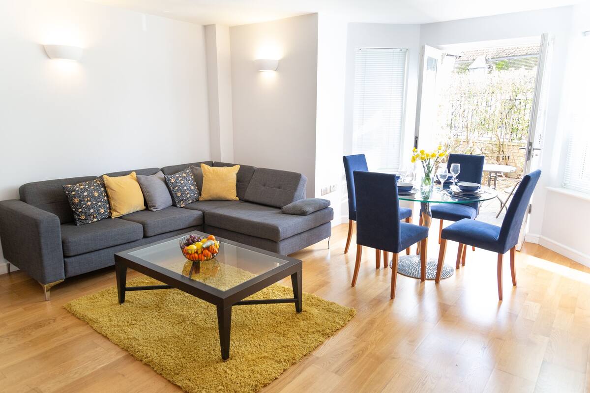 Een moderne woonkamer met een grijze modulaire bank, een glazen salontafel, en een eetgedeelte met blauwe stoelen en een glazen tafel.