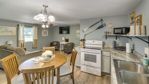 Eine kompakte Küche mit Mikrowelle, Herd und Spüle, ein Essbereich mit Holztisch und Stühlen sowie ein Wohnzimmer mit Sofa und Fernseher.