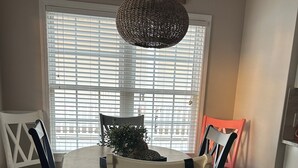 Un comedor con una mesa redonda, cuatro sillas, una lámpara colgante y una ventana con persianas.
