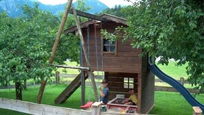 Una casita de juegos rústica de madera con un tobogán, rodeada por una cerca de madera y árboles.