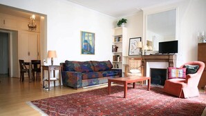Un salon comprenant un canapé, un fauteuil, une table basse et une cheminée. On y trouve également une bibliothèque et un téléviseur.
