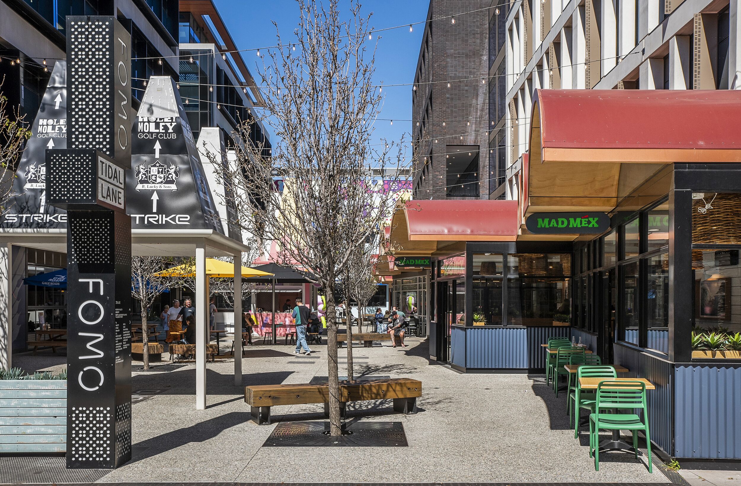 A street with outdoor seating, a tree, and various storefronts including a golf club and a restaurant.