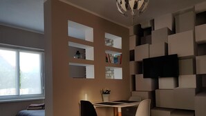 Un salon moderne comprenant un coin repas, un téléviseur à écran plat fixé sur un mur aux motifs géométriques, une fenêtre offrant une vue agréable et un coin salon confortable avec un lit.