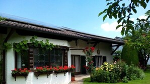 Un bâtiment blanc avec un toit recouvert de tuiles, de grandes fenêtres dotées de volets verts et un jardin bien entretenu où poussent diverses plantes et fleurs.