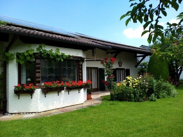 A white building with a tiled roof, large windows with green shutters, and a well-maintained garden with various plants and flowers.