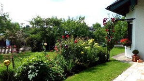 Un jardin avec différentes plantes et fleurs, un sentier et un bâtiment à l’extérieur blanc et au toit foncé.